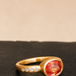 A gold ring featuring a large oval orange-red gemstone and a row of small clear stones on the band, resting on a dark textured surface against a warm beige background.