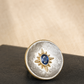 A round silver-toned ring with a textured finish, featuring a gold-toned sunburst motif in the center enclosing a small oval blue gemstone. The ring is displayed on a dark textured surface against a warm beige background.