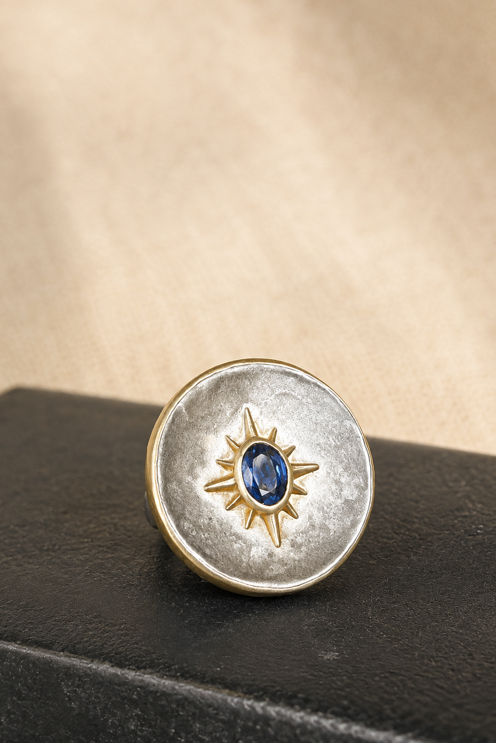 A round silver-toned ring with a textured finish, featuring a gold-toned sunburst motif in the center enclosing a small oval blue gemstone. The ring is displayed on a dark textured surface against a warm beige background.