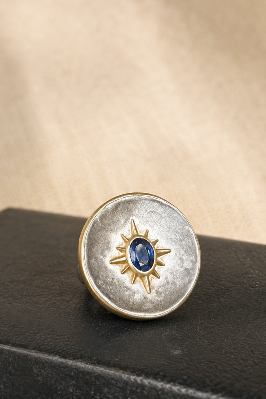 A round silver-toned ring with a textured finish, featuring a gold-toned sunburst motif in the center enclosing a small oval blue gemstone. The ring is displayed on a dark textured surface against a warm beige background.